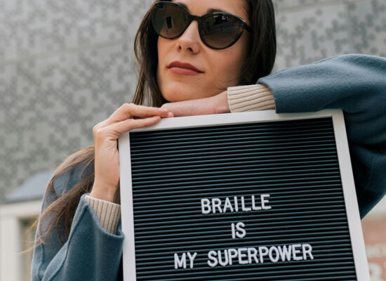 Eine Frau mit braunen langen Haaren lehnt sich auf ein Schild auf dem „BRAILLE IS MY SUPERPOWER“ steht. Sie hat einen blauen Mantel an und trägt eine dunkle Brille.