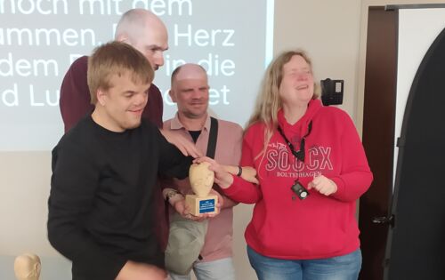 Bei der Preisverleihung nehmen Lucas Brinse, Reiner Delgado und Annette Sander den Preis entgegen. Sie halten die Skulptur gemeinsam in den Händen: Das Herz aus hellem Holz, auf dem ein Auge und ein Ohr zu sehen sind, steht auf einem Sockel.