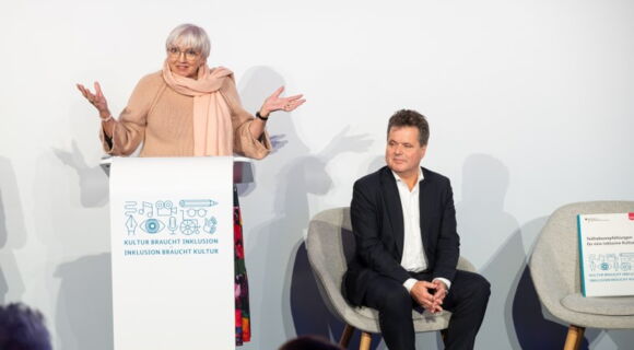 Claudia Roth, Ministerin für Kultur und Medien, steht mit ausgebreiteten Armen an einem Rednerpult, darauf ein Logo mit dem Text: „Kultur braucht Inklusion, Inklusion braucht Kultur“. Daneben sitzt der Bundesbehindertenbeauftragte, Jürgen Dusel.