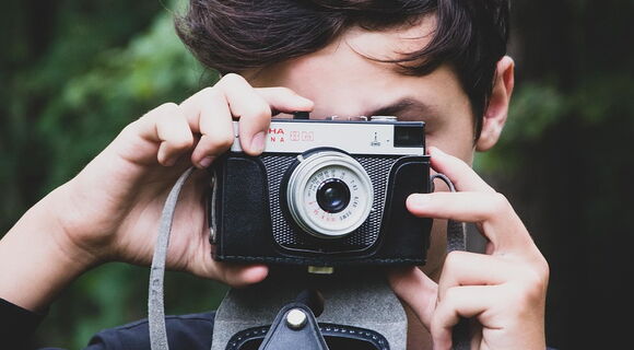 Ein junger Mensch mit dunklen Haaren hält eine analoge Kamera vor sein Gesicht und fotografiert. Die Kamera hat ein Vintage-Design mit schwarzem Gehäuse und silbernen Details. Der Hintergrund ist grün und unscharf.