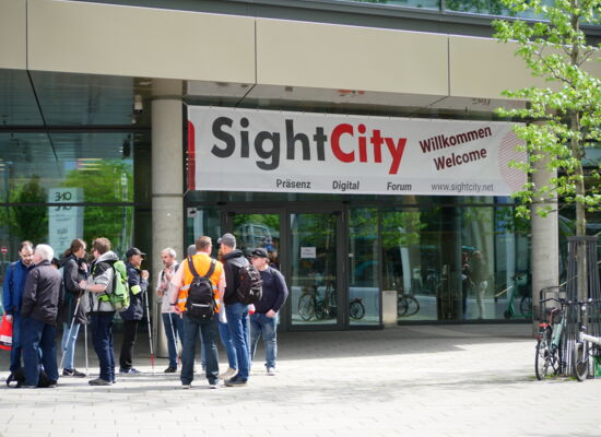 : Vor dem Eingang der Sightcity steht eine Gruppe von Menschen. Viele von Ihnen haben einen Langstock dabei. Sie tragen Rucksäcke. Über der Tür hängt ein Willkommens-Banner.