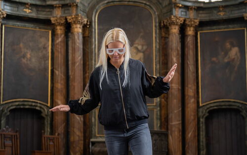 Eine junge Frau mit Simulationsbrille geht in einer Kirche drei nicht markierte Stufen hinunter. Unsicher balanciert sie mit den Armen.