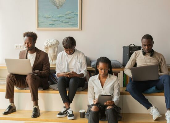 Mit unterschiedlichen Geräten am selben Projekt arbeiten: Vier junge Menschen sitzen nebeneinander auf einer breiten Bank und gucken jeweils in einen Laptop, ein Tablet oder ein Handy. Es sind vier junge Personen in schlichter, moderner Kleidung.