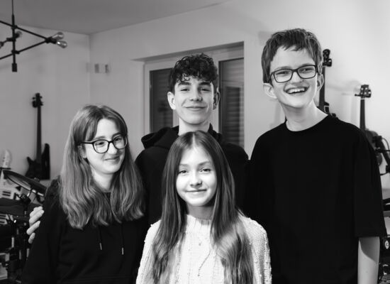 Die Band Rockytrio, vier Jugendliche zwischen 13 und 16, stehen in einem Musikraum vor Instrumenten und schauen freundlich lächelnd in die Kamera.