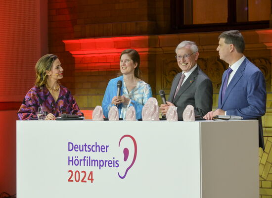 Auf der Bühne hinter einem weißen Pult mit dem Logo des Deutschen Hörfilmpreises stehen folgende Personen von links nach rechts: Nadine Heidenreich, Dr. Ulrike Köhler, Bundespräsident a. D. Prof. Dr. Horst Köhler und Dr. Thomas Krämer