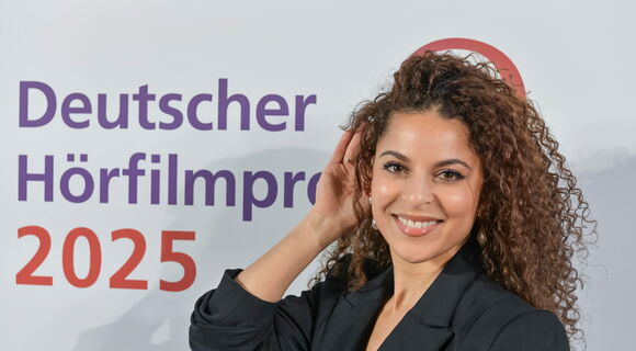 Patricia Meeden, eine jüngere Frau mit strahlendem Lächeln und langen, braunen Locken, hält eine Hand ans Ohr. Auf der Wand hinter ihr steht "Deutscher Hörfilmpreis 2025"