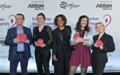 Beim Deutschen Hörfilmpreis steht ein AD-Team steht vor einer Sponsorenwand. Sie halten die Trophäe ADele in ihren Händen. Alle fünf Personen lächeln breit.