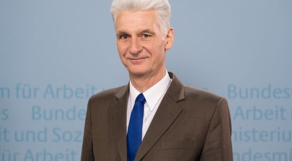 Rolf Schmachtenberg hat weißes, zurückgekämmtes Haar. Er trägt einen braunen Anzug, ein weißes Hemd und eine blaue Krawatte. Im Hintergrund ist unscharf der Text: „Bundesministerium für Arbeit und Soziales“ angedeutet.