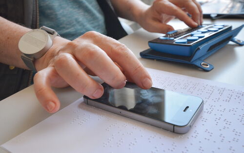 Zwei Hände sind zu sehen: Die linke bedient eine Braillezeile, die rechte ein Smartphone. Das Smartphone liegt auf einer mit Brailleschrift bedruckten Seite.