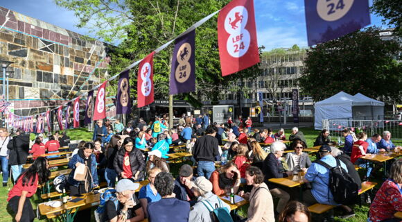 Berliner Platz neben der Liederhalle. Auf einer Rasenfläche stehen im Sonnenlicht Bierbänke und Tische, an denen zahlreiche Menschen sitzen. Über ihnen ist eine Leine gespannt, an der Fähnchen mit dem Logo des Louis Braille Festivals hängen.