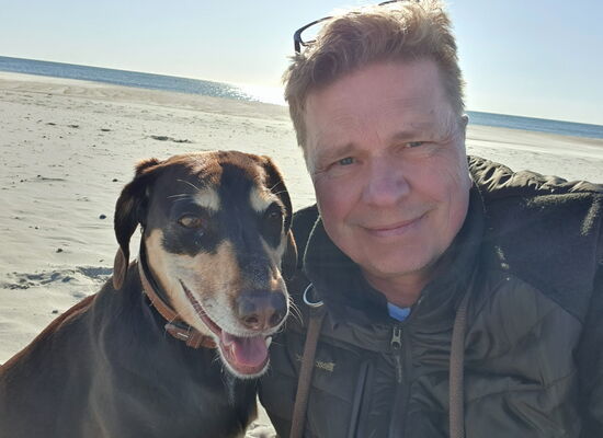 Ein Mann mit kurzen, hellen Haaren und einer dunklen Jacke macht ein Selfie mit einem braun-schwarzen Hund am Sandstrand. Beide schauen freundlich in die Kamera. Im Hintergrund sind Meer, blauer Himmel und Sonnenlicht zu sehen.