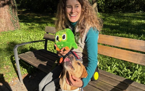 Vivian Hösch sitzt lächelnd im Sonnenschein auf einer Bank. In ihrer rechten Hand hält sie Duo, eine fußballgroße grüne Plüscheule, das Maskottchen von Duolingo. Mit Links streichelt sie ihren hellbraunen Blindenführhund Candy, der neben ihr sitzt.