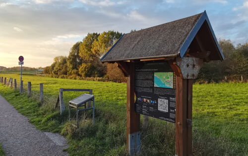 Am Wegesrand steht eine Infotafel zum Planeten Erde, daneben ist die entsprechende Station des Planetenlehrpfades in Wedel. Der Weg führt weiter in die Ferne, umgeben von Wiesen.