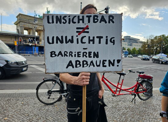 Ein Mann hält ein Schild in die Kamera: „UNSICHTBAR ≠ UNWICHTIG – BARRIEREN ABBAUEN“. Er steht vor dem Brandenburger Tor. Im Hintergrund ist ein rotes Tandem zu sehen.
