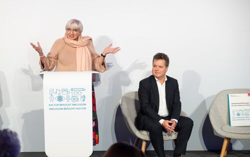 Claudia Roth, Ministerin für Kultur und Medien, steht mit ausgebreiteten Armen an einem Rednerpult, darauf ein Logo mit dem Text: „Kultur braucht Inklusion, Inklusion braucht Kultur“. Daneben sitzt der Bundesbehindertenbeauftragte, Jürgen Dusel.