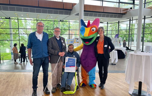 Gruppenbild: Drei Männer, einer davon im Rollstuhl, eine Frau und das Kampagnen-Maskottchen, ein Chamäleon, stehen in einer Empfangshalle. Heinrich Schaudt, Vorsitzender Arbeitsgemeinschaft Barrierefreiheit Rhein-Neckar, präsentiert die Gewinner-Urkunde.