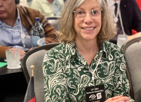 Sabine Ström sitzt während der Generalversammlung an einem Tisch mit Deutschland-Fähnchen und dem Schild „Germany“. Sie trägt mittellanges helles Haar, eine Brille und ein Kleid mit floralen Mustern. Hinter ihr sind weitere Teilnehmer zu erkennen.