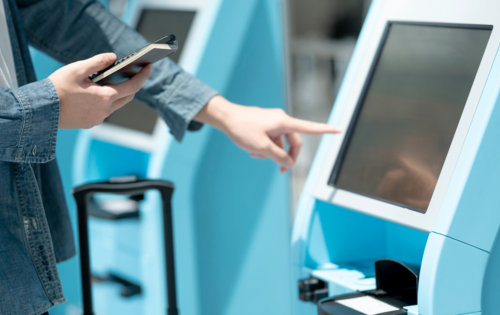 Jemand steht vor einem Automaten und zeigt mit dem Finger darauf. In der anderen Hand hält er ein Handy und ein kleines Heft.