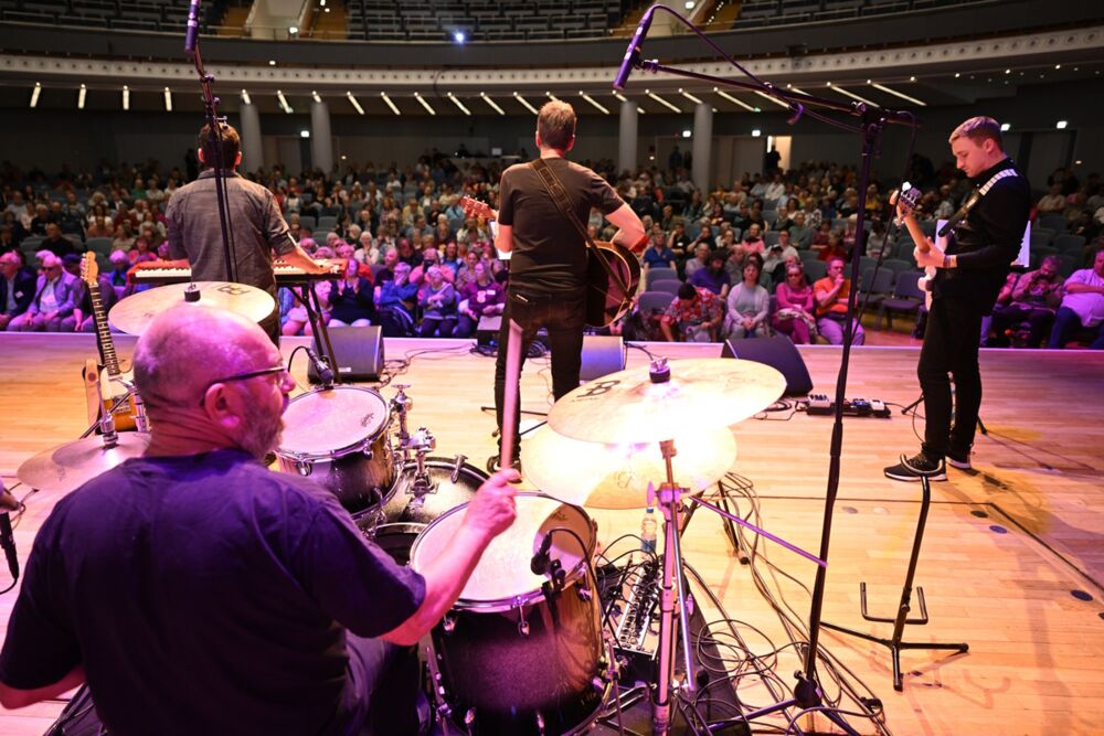 Der Coverband Warehouse über die Schulter ins Publikum fotografiert: Die Musiker spielen Schlagzeug, Gitarre und Keyboard.