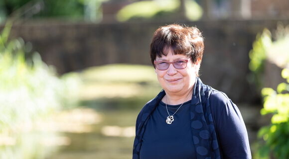 Margit Giegerich trägt kurzes dunkles Haar, eine Brille, dunkle Kleidung und eine Kette mit blütenförmigem Anhänger. Sie steht auf einem sonnigen Weg, im Hintergrund sind verschwommen Büsche und Bäume zu sehen.