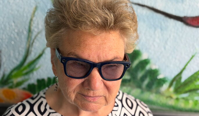 Dagmar Krause ist in Nahaufnahme vor einer farbig gestalteten Wand zu sehen. Sie trägt eine dunkle Brille und ein gemustertes Oberteil.