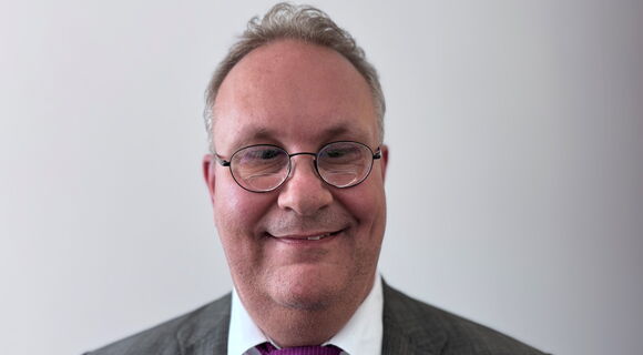 Joachim Günzel lächelt in die Kamera. Er trägt ein dunkelgraues Sakko über einem weißen Hemd, kombiniert mit einer beerenfarbenen Krawatte. Dazu eine unauffällige Brille mit runden Gläsern.