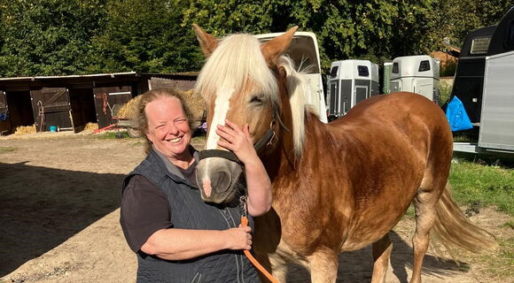 Linda Pröve steht bei einem Haflingerpony, dessen Kopf sie mit einer Hand umfasst. Ihr langes lockiges Haar trägt sie zusammengebunden. Im Hintergrund sind Bäume und Pferdeanhänger zu sehen.