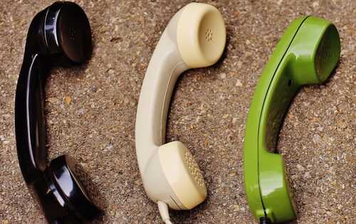 Drei klassische Telefonhörer in Schwarz, Beige und Grün liegen nebeneinander auf einem Korkuntergrund.