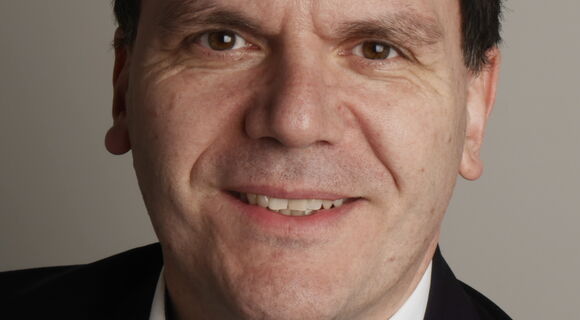 Prof. Dr. Thomas Ach blickt lächelnd in die Kamera. Er hat dunkles kurzes volles Haar mit geradem Ansatz. Er trägt ein schwarzes Sakko und ein weißes Hemd.