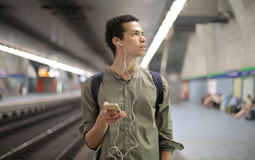 Ein Mann mit dunklen, krausen Haaren steht mit einem Handy in der Hand und Kopfhörern in den Ohren auf einem Bahnsteig. Er blickt mit gerunzelter Stirn irritiert zur Seite. Er trägt ein grünes lockeres Hemd.