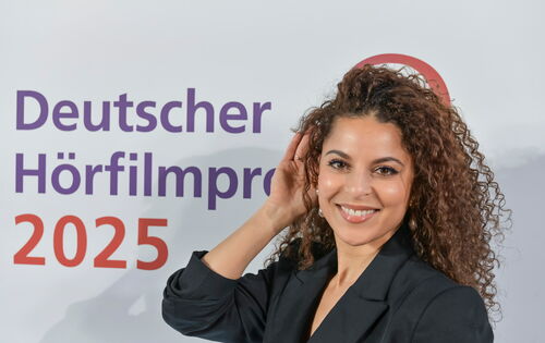 Patricia Meeden, eine jüngere Frau mit strahlendem Lächeln und langen, braunen Locken, hält eine Hand ans Ohr. Auf der Wand hinter ihr steht "Deutscher Hörfilmpreis 2025"