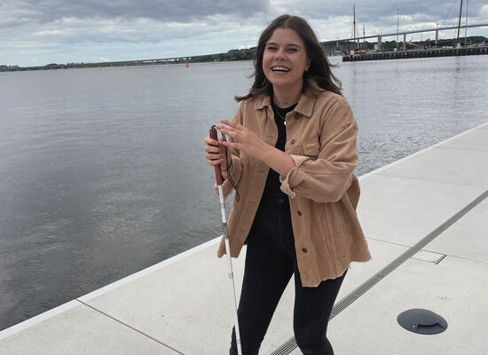 Jacqueline Eschmann steht an einer Hafenkante, dahinter befindet sich ein größeres Gewässer; im Hintergrund sind Schiffe und eine Brücke zu sehen. Sie hat langes dunkles Haar, trägt eine dunkle Hose, weiße Sneaker und hält ihren Langstock fest.