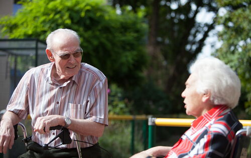 Ein alter Mann und eine alte Frau im Gespräch: Er trägt ein gestreiftes Hemd und stützt sich auf seinen Rollator. Sie sitzt rechts von ihm in einer rot-schwarz-karierten Bluse. Sie sitzen draußen, es ist sonnig.