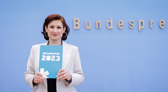 Die Antidiskriminierungsbeauftragte der Bundesregierung, Ferda Ataman, hält den Jahresbericht ihrer Einrichtung in den Händen. Sie hat nackenlanges dunkles Haar, trägt große Kreolen und einen weißen Blazer.