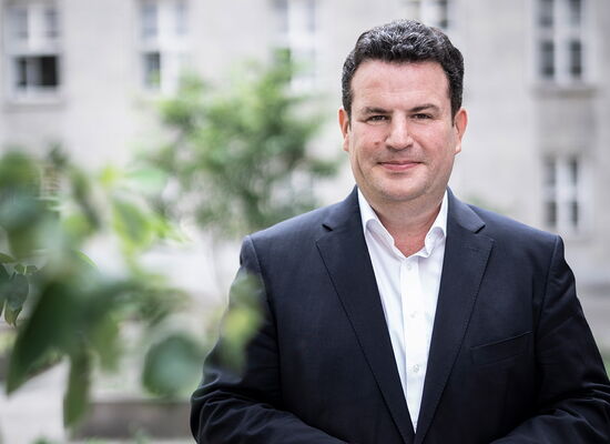 Bundesarbeitsminister Hubertus Heil schaut lächelnd direkt in die Kamera. Er trägt einen dunklen Sakko mit einem weißen Hemd. Er hat schwarze Haare, sein Bart ist kurz rasiert. Im Hintergrund sind helle Gebäude und Pflanzen zu sehen.
