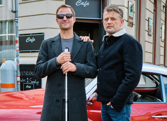Das Foto zeigt Philipp Hochmair als Alex Haller mit Sonnenbrille und Langstock sowie Andreas Guenther als Niko Falk, der ihm eine Hand auf die Schulter legt.
