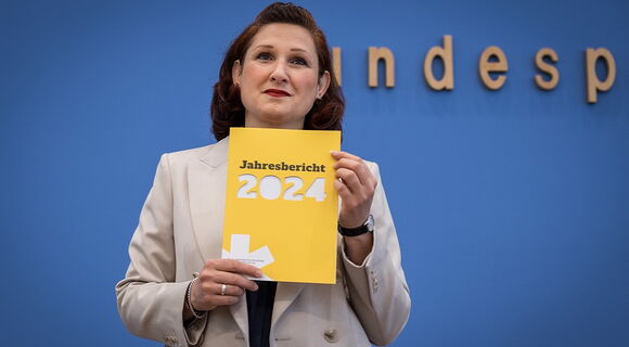 Das gelbe Heft mit dem Jahresbericht der Antidiskriminierungsstelle hält Ferda Ataman bei der Bundespressekonerenz hoch. Die unabhängigen Bundesbeauftragte hat braune, ondulierte Haare und trägt einen hellen Blazer.