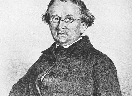 Zeichnung eines Mannes, Eduard Mörike, mit Brille und gewelltem Haar. Er trägt einen dunklen Mantel mit mehreren Knöpfen und sitzt mit leicht geneigtem Kopf. Der Hintergrund ist schlicht.