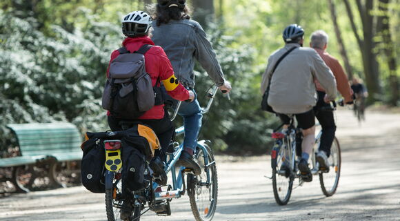 Zwei Tandems fahren hinter einander durch einen sonnigen Park. Sie sind mit Fahrradtaschen und Rucksack bepackt. Um den Arm einer Radfahrerin und hinten am Fahrrad ist die Blindenplakette beschäftigt. Sie tragen lockere Kleidung und Fahrradhelme.