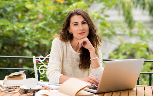 Eine Frau mit langen braunen Haaren sitzt draußen an einem Tisch. Im Hintergrund sind verschwommen Bäume zu sehen. Vor ihr liegen mehrere Bücher und ein Laptop. Die Hand unter dem Kinn haltend, blickt sie versonnen in die Kamera.
