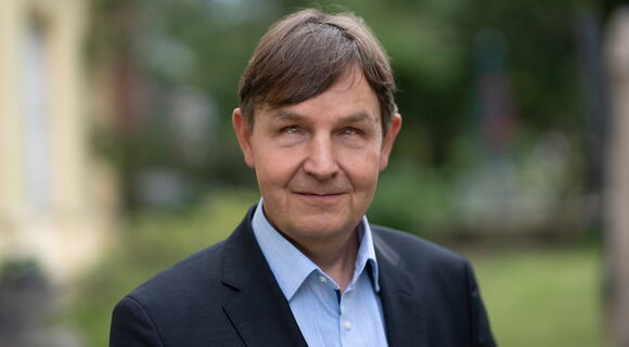 Ein Mann mittleren Alters mit kurzen, braunen Haaren trägt ein hellblaues Hemd und ein dunkelblaues Sakko. Er schaut leicht nach oben und lächelt dezent. Der Hintergrund ist unscharf und zeigt eine grüne, natürliche Umgebung.