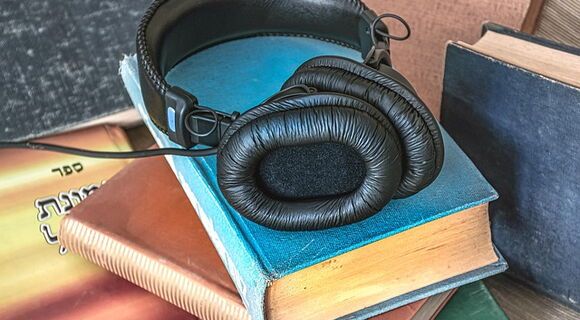 Ein schwarzer Kopfhörer mit gepolsterten Ohrmuscheln liegt auf einem Stapel Bücher. Auch daneben stehen Bücher.