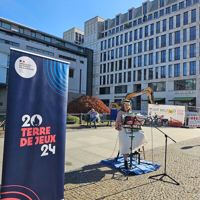 Zur Verabschiedung singt die Künstlerin Annika von Lier Lieder auf Französisch und Deutsch zum Akkordeon. Mit weißem Faltenrock und einem rot-weiß-gestreiften Oberteil sitzt sie neben einem blauen Aufsteller mit der Aufschrift: „20 - Terre de Jeux - 24“.