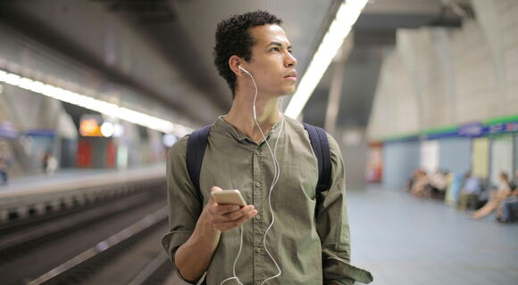 Ein Mann mit dunklen, krausen Haaren steht mit einem Handy in der Hand und Kopfhörern in den Ohren auf einem Bahnsteig. Er blickt mit gerunzelter Stirn irritiert zur Seite. Er trägt ein grünes lockeres Hemd.