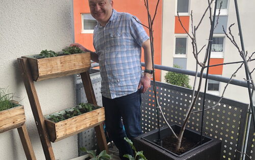 Rainer Heim trägt ein helles, kariertes Hemd und steht lächelnd auf seinem Balkon. Neben ihm ein dreistufiges Hochbeet mit Kräutern, vor ihm einige Blumenkübel mit wachsenden Pflanzen.