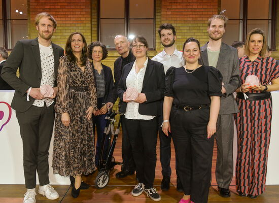 In festlicher Kleidung steht das AD-Teamder Drama-Miniserie „Deutsches Haus“  auf der Bühne. Es gewann die ADele in der Kategorie „Serie“. Einige Team-Mtglieder halten die Keramik-Trophäe in den Händen.