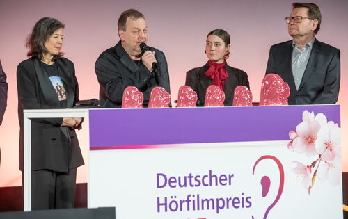 Klaus-Jörg Kaminski, blinder AD-Autor des Films „Paris, Texas“ spricht in ein Mikrofon bei der Deutschen Hörfilm-Verleihung. Neben ihm stehen sein Team und die Laudatorin des Films. Auf einem Tisch stehen sechs rosafarbene Hörfilmpreis-Trophäen.