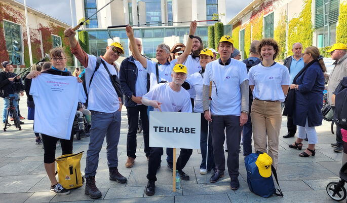 In weißen T-Shirts vom DBSV steht eine Gruppe von neun Menschen vor dem Bundeskanzleramt. Sie halten ein Schild mit der Aufschrift „Teilhabe“ und ihre Blindenstöcke nach oben.