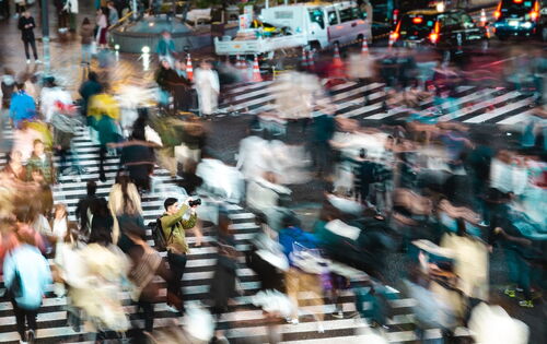Eine belebte Kreuzung: zahllose Menschen, verschwommen dargestellt, überqueren Überwege. Im Hintergrund, ebenfalls undeutlich, Fahrzeuge.