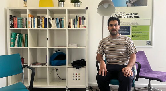 Saiman Mehra sitzt im Raum der Psychologischen Studienberatung. Er hat dunkles Haar und einen Vollbart. Hinter ihm an der Wand steht ein weißes Regal mit quadratischen Fächern, gefüllt mit Büchern und anderen Dingen.